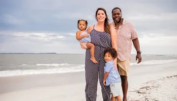 A happy family with two young children is standing on a beach, smiling and posing for a photo with the ocean in the background.