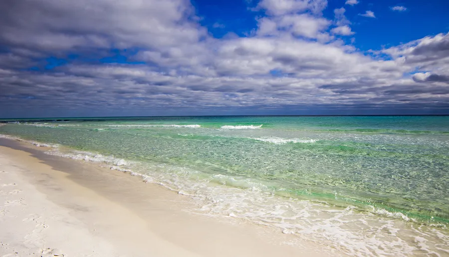 A serene beach scene with clear turquoise waters, white sands, and a sky partially covered with clouds.