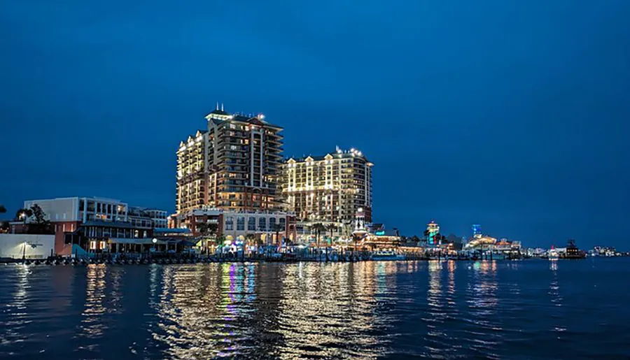 The image shows a vibrant evening view of a waterfront cityscape with illuminated buildings and lights reflecting on the water's surface.