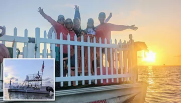 A group of people is posing happily behind a white picket fence on a waterfront at sunset.
