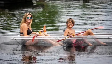 A smiling woman and a young boy are sharing a clear kayak, both holding paddles, on a calm water body.