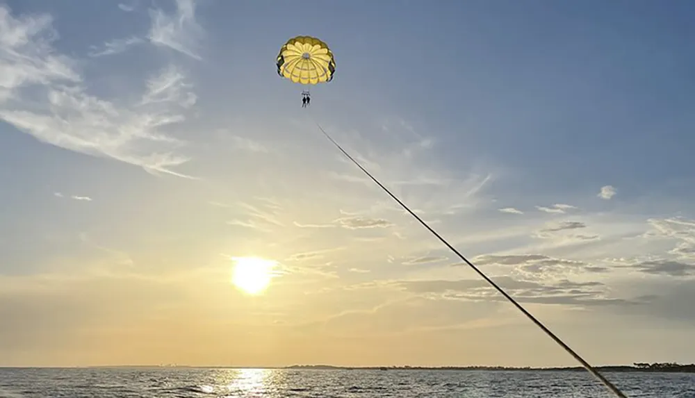 A person is parasailing over the ocean during sunset attached to a boat by a long tether