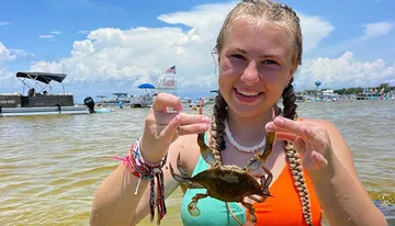 A smiling young woman is holding up a crab at a sunny waterfront setting, with boats in the background.