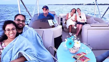 A group of people are enjoying a boat ride, with a couple snuggled under a blanket in the foreground and two women sitting with snacks at a table, while a man pilots the boat in the background.