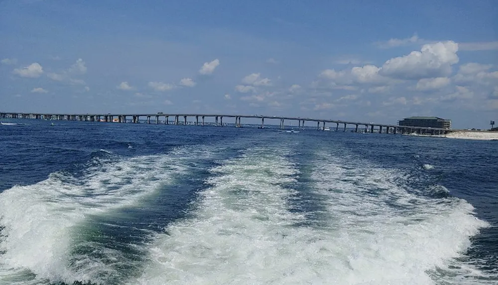 A boat leaves frothy white waves in its wake with a long bridge spanning the background under a partially cloudy sky