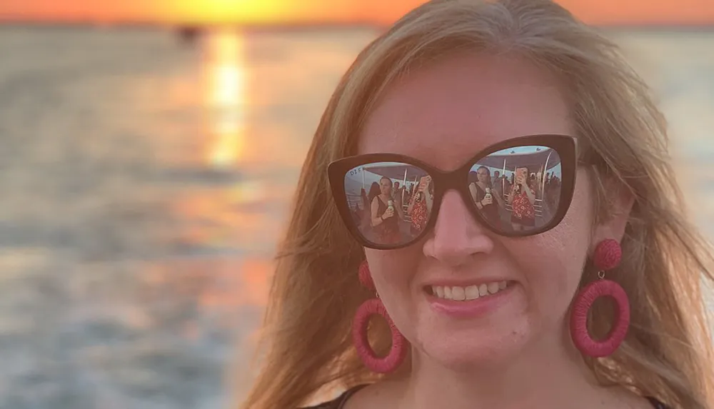 A smiling person at sunset with the reflection of people in her sunglasses