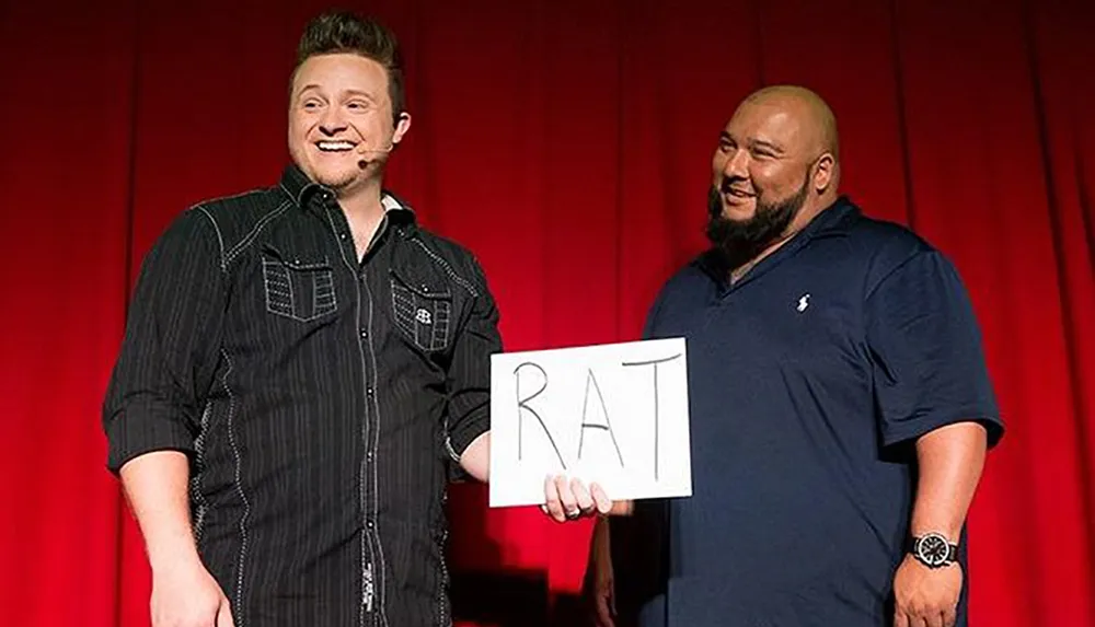 Two men are smiling on stage one of whom is holding a sign with the word RAT written on it possibly during a comedic or entertainment performance