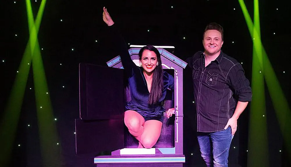 A smiling woman sits in a magic assistants box with her body apparently divided while a male magician stands beside her set against a backdrop of green stage lights