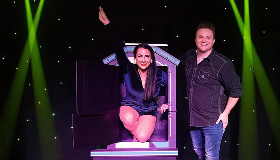 A smiling woman sits in a magic assistant's box with her body apparently divided, while a male magician stands beside her, set against a backdrop of green stage lights.