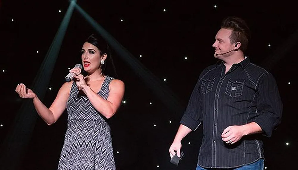A woman is singing into a microphone while a man stands beside her both on a stage with a starry background and spotlights shining down