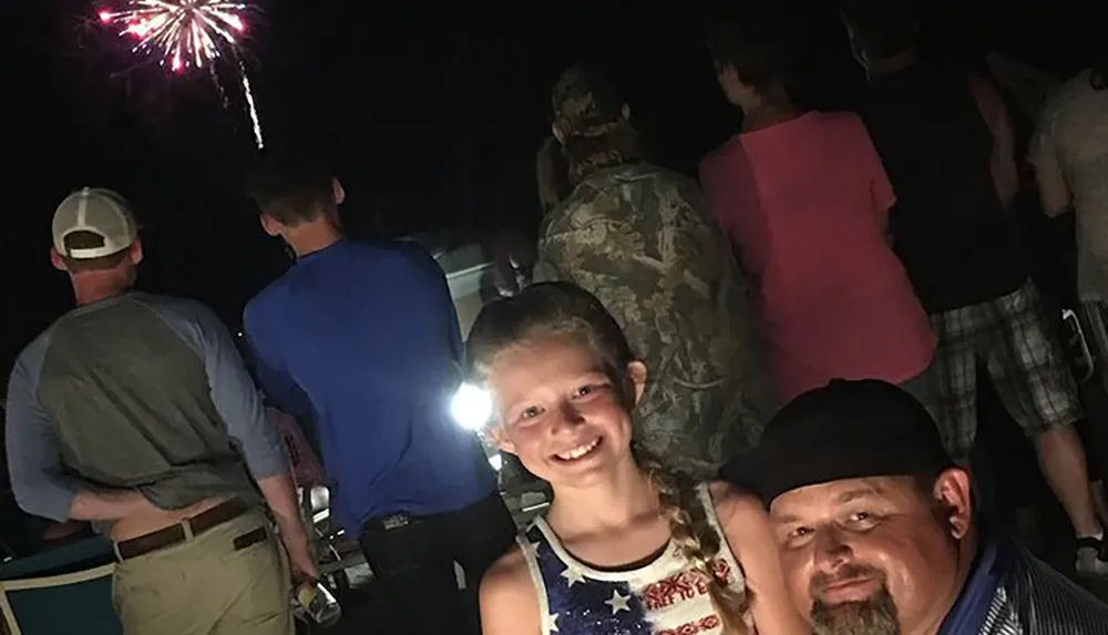 A girl smiles for the camera with a man beside her while other people watch fireworks at night