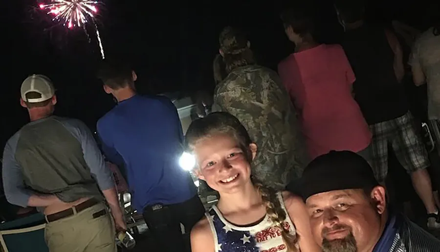 A girl smiles for the camera with a man beside her while other people watch fireworks at night.