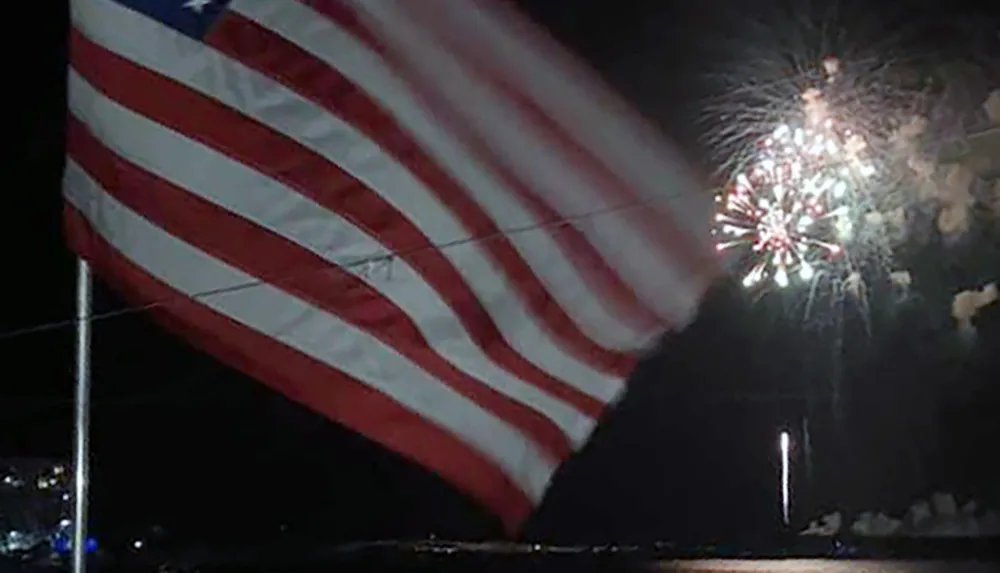 An American flag is prominently displayed in the foreground with fireworks bursting in the night sky behind it