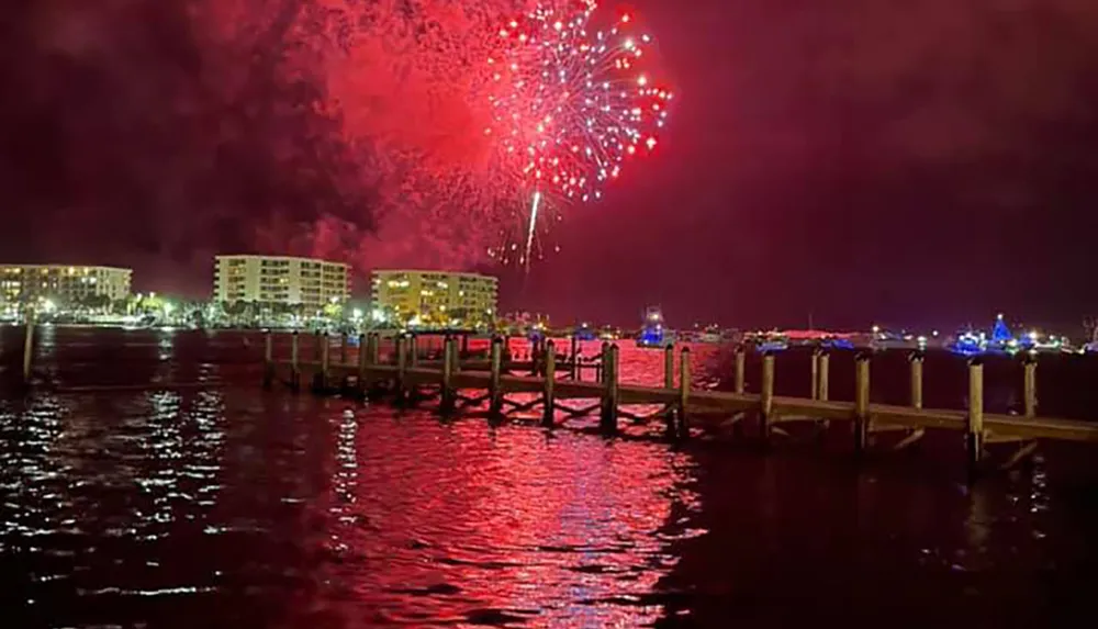 The image shows a vibrant fireworks display lighting up the night sky over a body of water with reflections on the water and a pier in the foreground against the backdrop of urban buildings