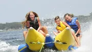 Three children are joyously riding a yellow inflatable banana boat on a sunny day, splashing through the water.