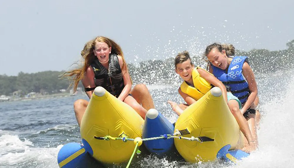 Three children are joyously riding a yellow inflatable banana boat on a sunny day splashing through the water