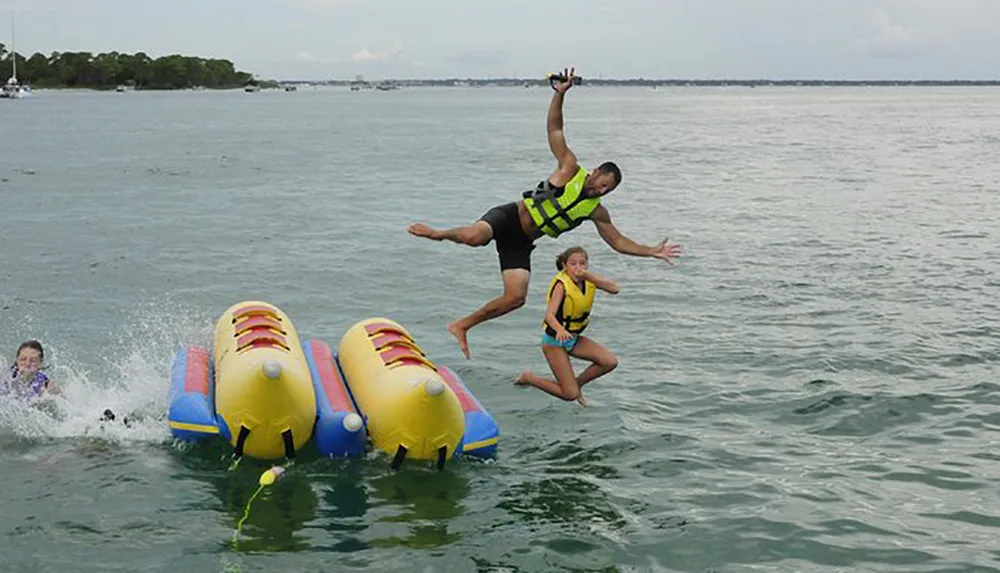 Two people are airborne over the water mid-jump from an inflatable water raft with one person already in the water and another on the raft
