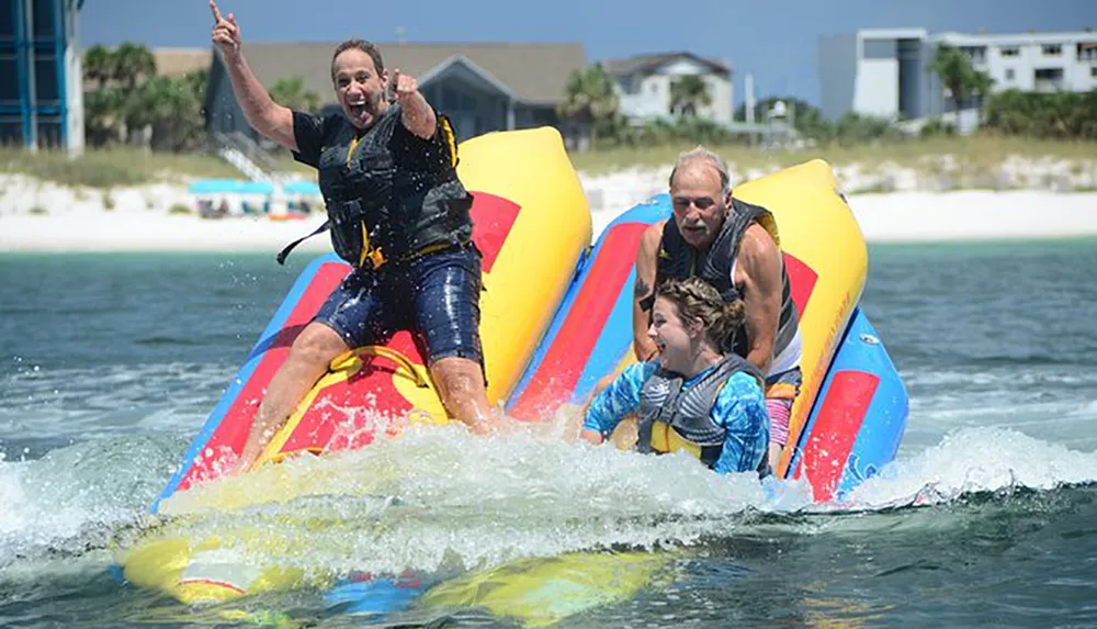 Three people are riding a colorful inflatable towable tube on a body of water with the person at the front raising their hand in excitement