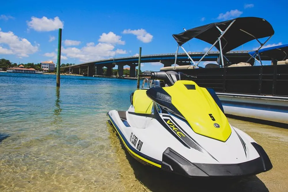 A yellow and white jet ski is docked near the shore with a pontoon boat beside it and a bridge in the background under a blue sky