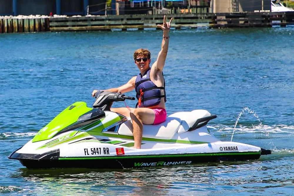 A person is joyfully raising their hand in a peace sign while riding a green and white Yamaha WaveRunner on a sunny day