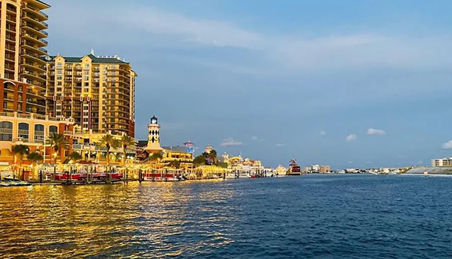 The image shows a picturesque waterfront with a blend of modern buildings and a lighthouse, against a blue sky dotted with clouds.