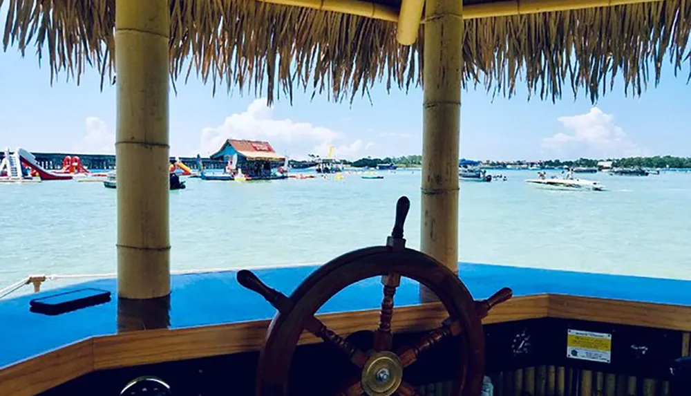 The image shows a view from a boat with a wooden steering wheel looking out over a lively tropical waterfront with other boats and structures framed by a thatched roof