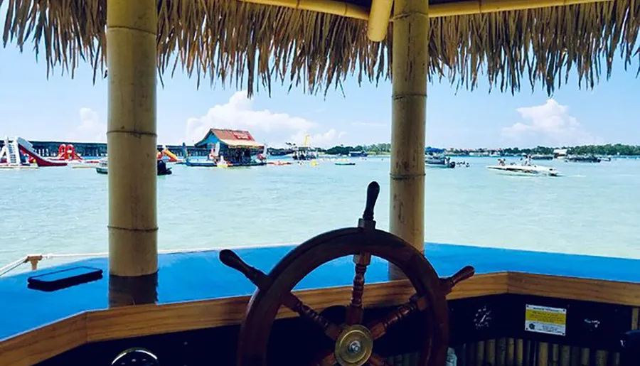 The image shows a view from a boat with a wooden steering wheel, looking out over a lively tropical waterfront with other boats and structures, framed by a thatched roof.