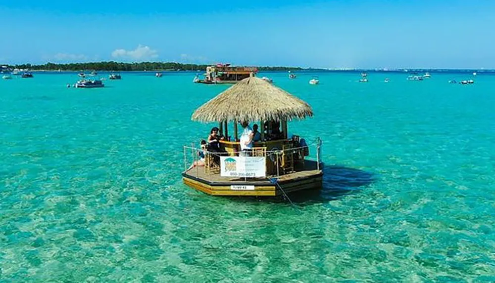 A small thatched hut bar floats on clear blue waters surrounded by various boats with people enjoying the sea