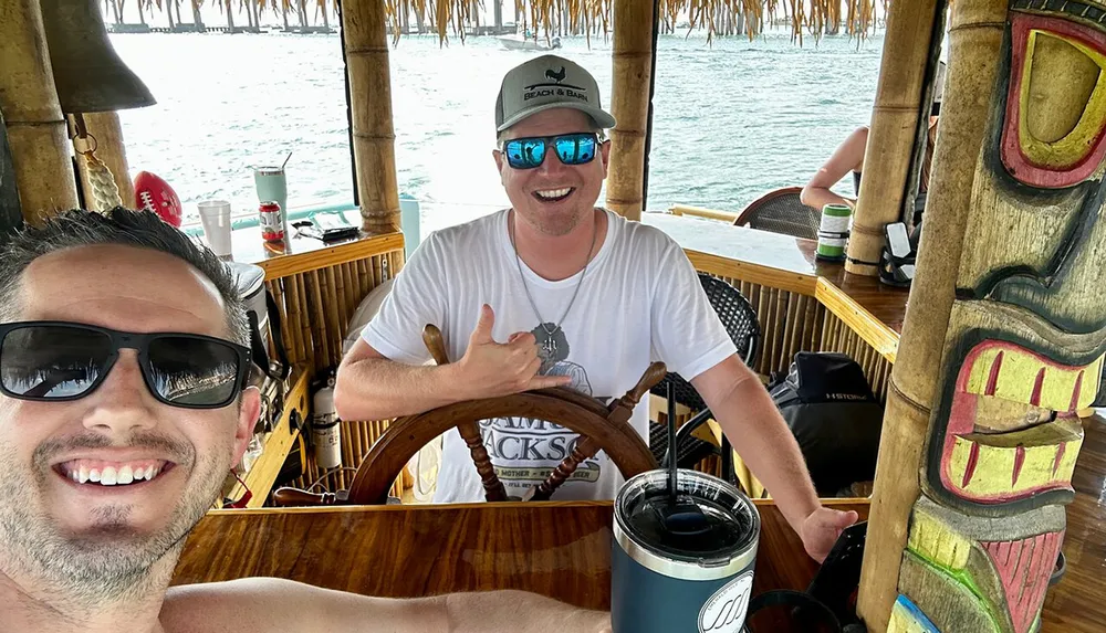 Two men are posing for a selfie at a bamboo-themed bar one holding the wheel and giving a thumbs-up with tropical vibes and the ocean in the background