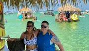 A man and a woman are smiling at the camera while posing under a tiki-style roof with a turquoise waterfront scene featuring floating platforms and people enjoying water activities in the background.