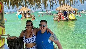 A man and a woman are smiling at the camera while posing under a tiki-style roof with a turquoise waterfront scene featuring floating platforms and people enjoying water activities in the background.