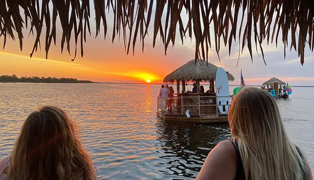 Two individuals are watching a vibrant sunset over the water where floating tiki bars create a festive atmosphere
