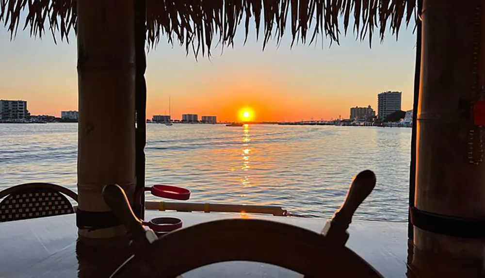 The image depicts a serene sunset view over the water from a thatched-roof structure framed by a bamboo pole and chair suggesting a tropical or beachside setting