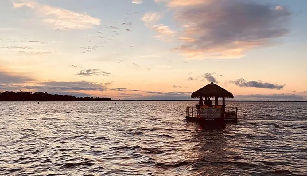 A thatched-roof structure atop a small floating platform sits on a body of water under a sunset sky