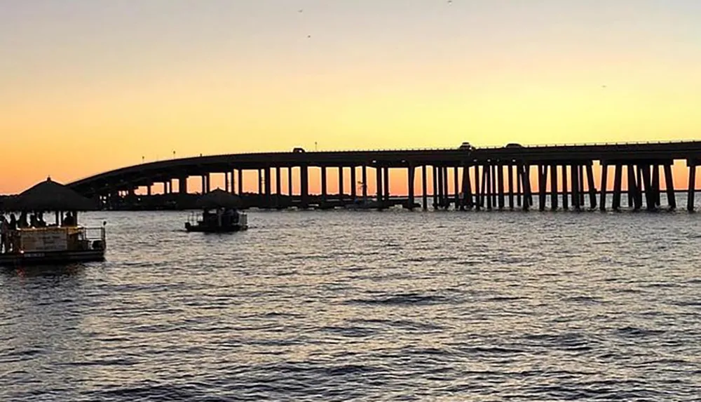 The image shows a long bridge extending over calm waters against a beautifully gradated sunset sky with silhouettes of boats on the water near the shore