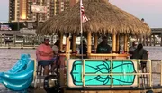 A group of people enjoys drinks on a floating tiki bar boat at dusk.