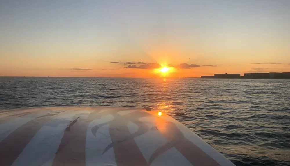 The image captures a serene sunset over the ocean viewed from the front of a boat with a striped pattern on its bow