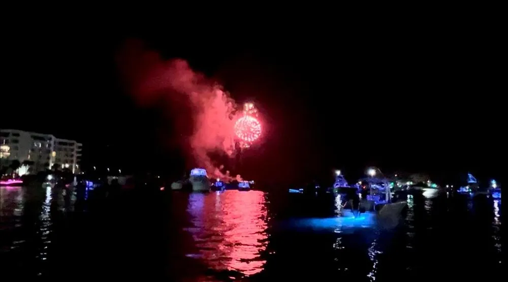 The image captures a festive night scene with vibrant fireworks in the sky over a body of water where boats illuminated with blue lights are scattered reflecting a serene and celebratory atmosphere