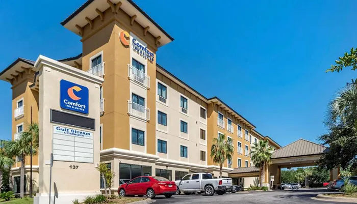 The image shows the exterior of a Comfort Inn  Suites hotel on a sunny day with cars parked in front and a partly visible sign for Gulf Stream Commons