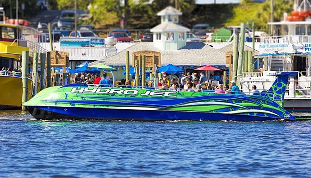 A vibrantly colored hydrojet boat is docked by a bustling waterfront with people and businesses in the background