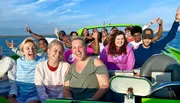 A group of joyful people are posing with excited expressions while seated on what appears to be a bright green boat, with a clear sky and water in the background.