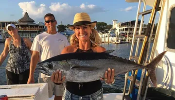 A smiling woman in a sun hat is proudly holding a large fish on a boat, with two other individuals standing behind her.