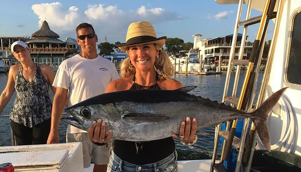 A smiling woman in a sun hat is proudly holding a large fish on a boat with two other individuals standing behind her