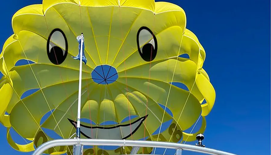 A large yellow parasail with a smiling face design is flying in the clear blue sky, attached to a boat by a harness.