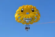 A group of people is parasailing with a bright yellow smiley-faced parachute against a clear blue sky.