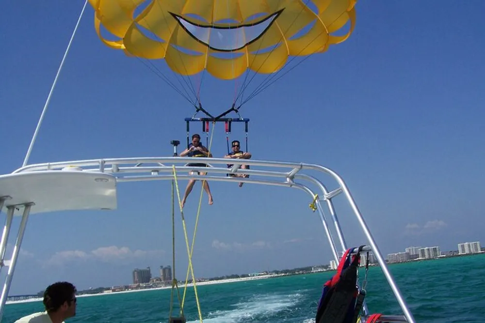 Two people are parasailing above the ocean being towed by a boat with a city skyline in the background