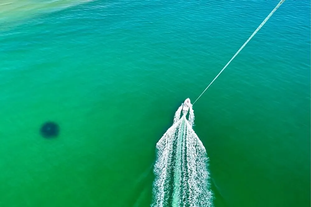 A birds-eye view of a boat cutting through green waters leaving a frothy wake as it tows a line or net
