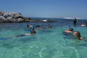 A group of people are snorkeling in clear, shallow waters near a rock formation.