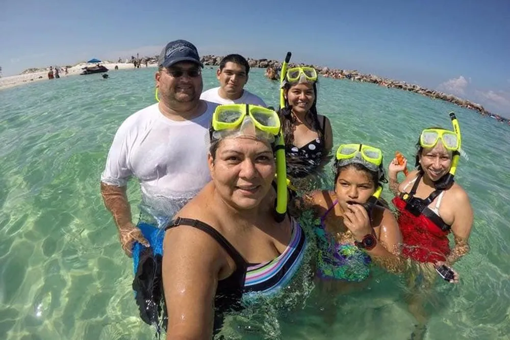 A group of people most wearing snorkeling gear are taking a selfie in clear shallow waters with a beach visible in the background