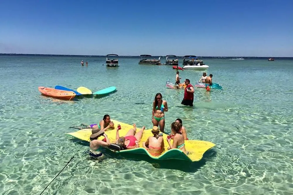 People are enjoying a sunny day on a clear and calm beach with some resting on a colorful floating mat and others wading or chatting in the shallow water surrounded by boats and personal watercraft
